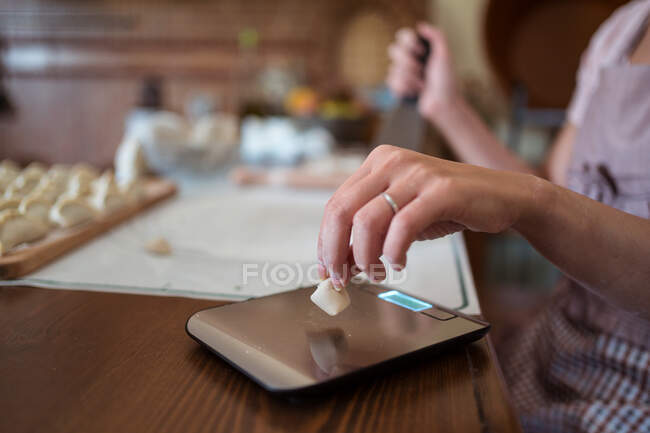 Cultivar cozinheira anônima pesando massa em balança eletrônica enquanto prepara jiaozi sentado à mesa na cozinha — Fotografia de Stock