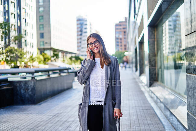 Emprendedora positiva vestida con ropa formal mientras habla por celular y mira hacia otro lado mientras sonríe. - foto de stock