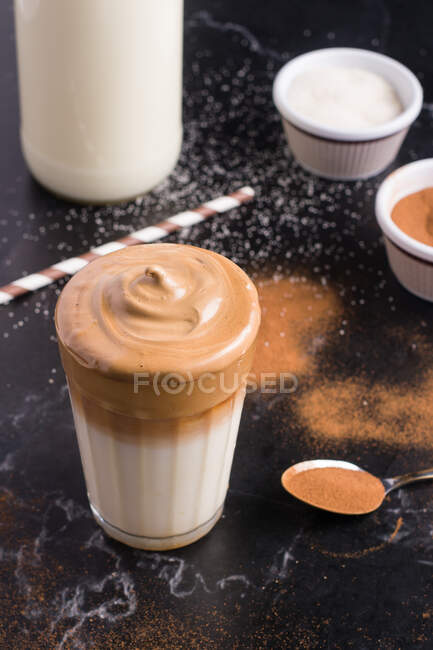 Bicchiere di delizioso caffè Dalgona con latte e condimento schiumoso posto su tavolo disordinato nero con cacao in polvere e zucchero — Foto stock