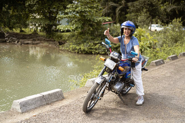 Full body of middle aged Asian female in casual clothes and protective helmet smiling and showing thumb up gesture while riding motorcycle near lake in tropical countryside — Stock Photo