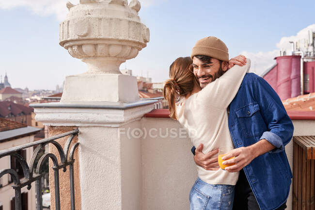 Боковой вид молодой девушки обнимающей парня, ярко улыбающейся стоя со стаканом сока на балконе — стоковое фото