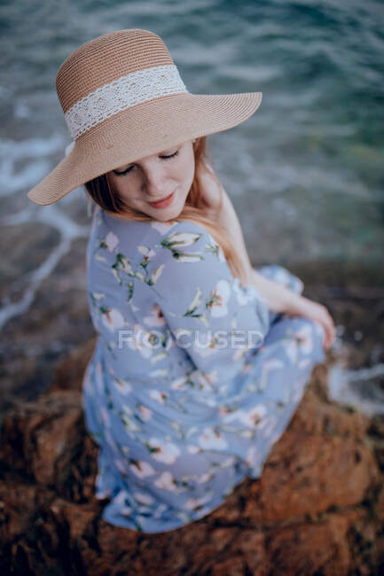Charming young female in summer dress and hat sitting on rocky seashore with closed eyes in summer evening — Stock Photo