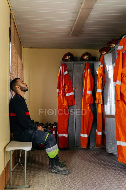 Vista lateral de cuerpo completo del joven bombero cansado en uniforme apoyado en la pared amarilla contra armarios con ropa protectora naranja colgando en las puertas dentro de la estación de bomberos - foto de stock