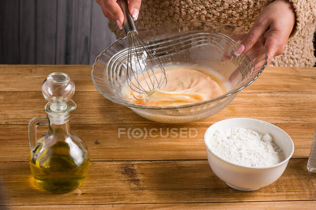 Crop anonymous female whisking eggs while preparing batter for crepes at wooden table with oil and flour in light kitchen — Stock Photo