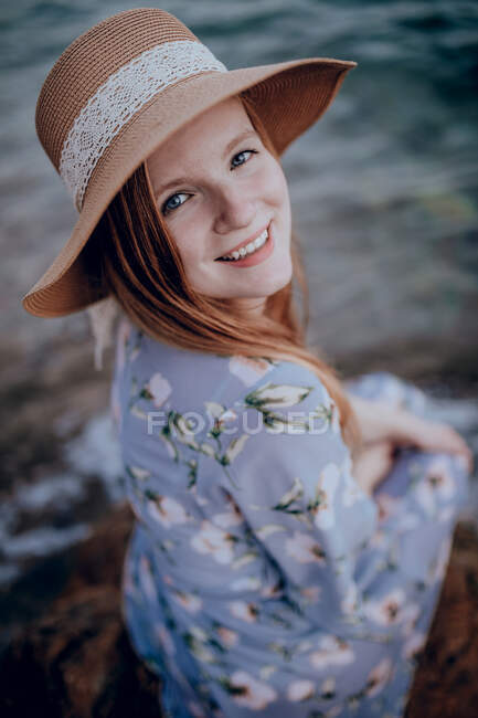 Encantadora joven hembra en vestido de verano y sombrero sentado en la costa rocosa mientras mira hacia otro lado en la noche de verano - foto de stock