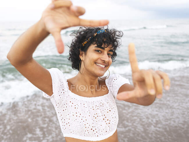 Charmante femme ethnique souriante montrant un panneau de cadrage debout sur la plage près de la mer en été et regardant la caméra — Photo de stock