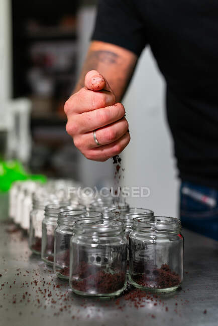 Crop male baker in casual outfit adding crumbs of chocolate cake into glass jars while preparing delicious layer dessert in kitchen — Stock Photo