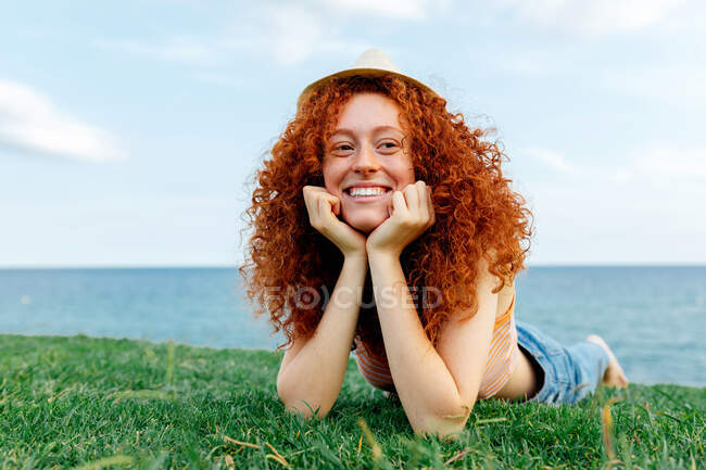 Щаслива кучерява руда жінка з веснянками лежить на газоні, дивлячись на узбережжя моря — стокове фото
