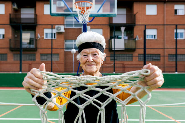 Старша жінка в спортивному одязі і головний убір дивиться на камеру, стоячи з сіткою в руці на баскетбольному майданчику з обручем — стокове фото