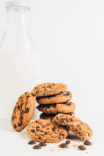 Tas de biscuits croquants sucrés avec des gouttes de chocolat placés sur la table avec bouteille vide dans la pièce lumineuse sur fond blanc — Photo de stock