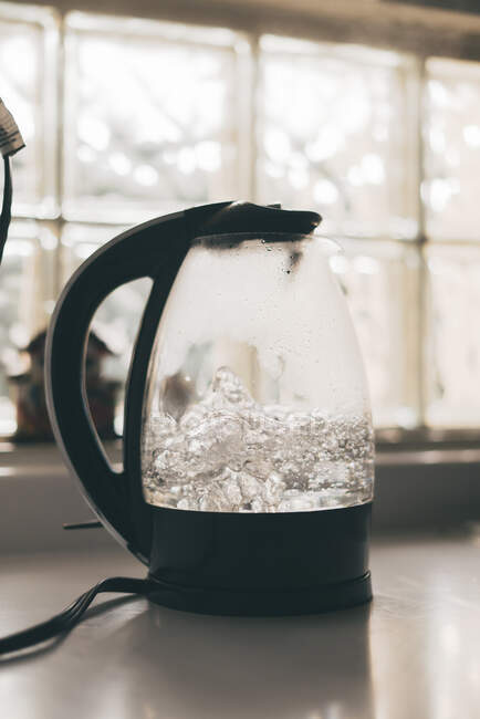 Transparent electric kettle with black handle and lid and with boiling water placed on white table in kitchen — Stock Photo