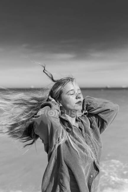 Black and white of young female in casual shirt standing on sandy beach near waving sea with eyes closed in summer — Stock Photo