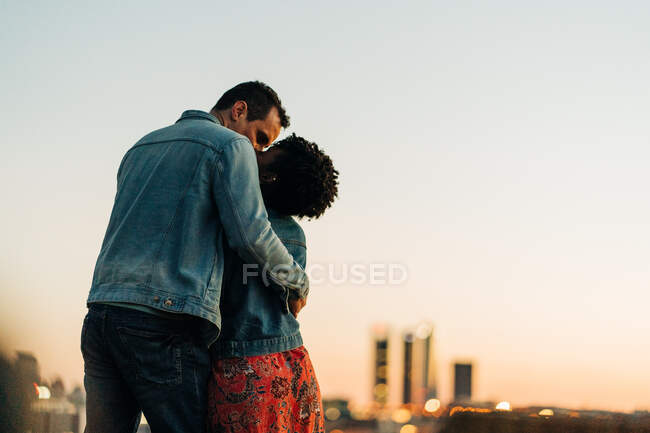 Романтична різноманітна пара обіймає і цілує, стоячи на газоні проти міського пейзажу з будівлями на розмитому фоні — стокове фото