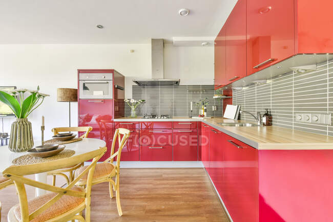 Современный интерьер кухни с красными шкафами и белым обеденным столом украшен цветами в вазе в современной квартире — стоковое фото