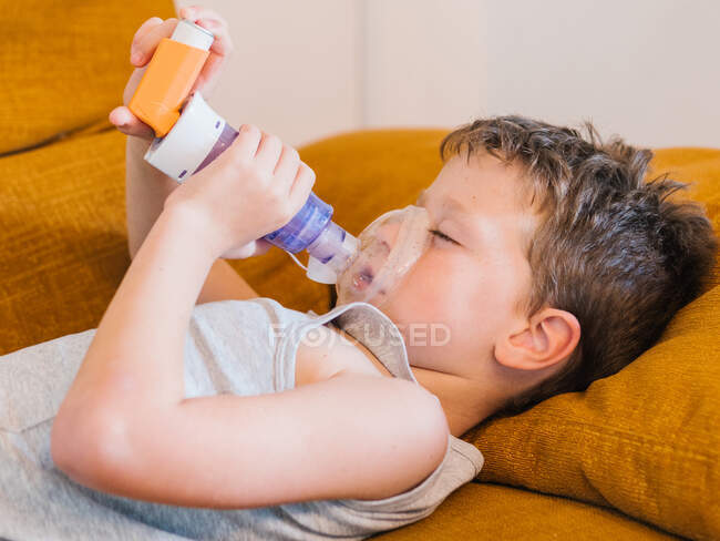 Vista laterale del bambino con asma usando l'inalatore mentre si sdraia sul divano a casa — Foto stock