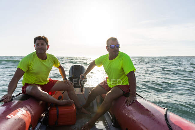 Longitud completa de salvavidas descalzos sentados en lancha motora inflable flotando en ondulante mar azul y controlando la seguridad en el mar - foto de stock