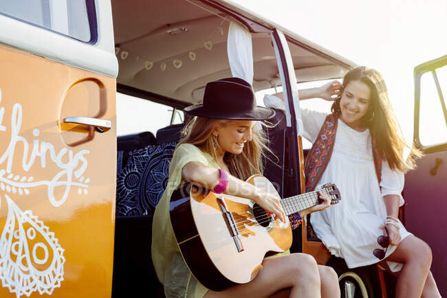 Two pretty girls dressed in summer clothes having fun outside a van and playing guitar — Stock Photo