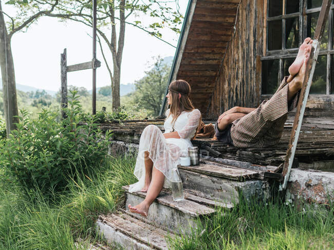 Привлекательная женщина в прозрачном платье смотрит в сторону, когда ее анонимный партнер спит со скрещенными руками и ногами на крыльце деревянного дома в сельской местности — стоковое фото
