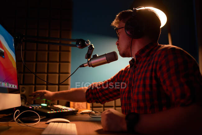 Вид збоку сфокусованого молодого чоловіка в картатій сорочці та окулярах, використовуючи комп'ютер та виступаючи в мікрофоні під час запису подкасту в темній студії — стокове фото