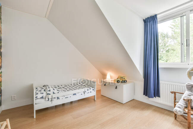 Дитяче ліжко розміщене біля білої шафи з іграшковим автомобілем у світлій просторій спальні з синьою завісою на вікні в квартирі — стокове фото