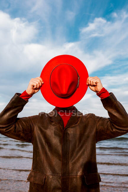 Personne méconnaissable en chapeau rouge et manteau debout près du rivage avec des pierres concassées en été — Photo de stock