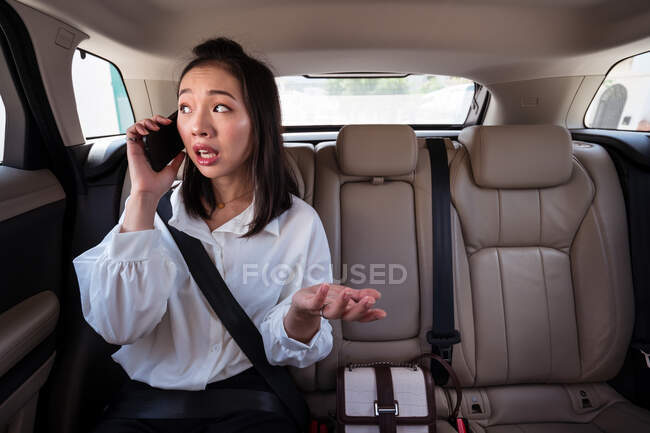 Passageiro étnico confuso com cinto de segurança apertado falando no celular enquanto andava no banco de trás em táxi — Fotografia de Stock