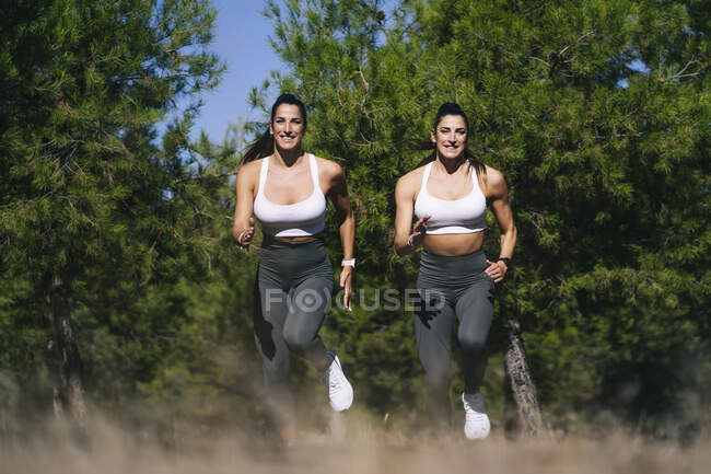 Cuerpo completo de gemelas deportivas sonrientes en ropa deportiva corriendo juntas en el campo durante el entrenamiento de fitness contra árboles verdes - foto de stock