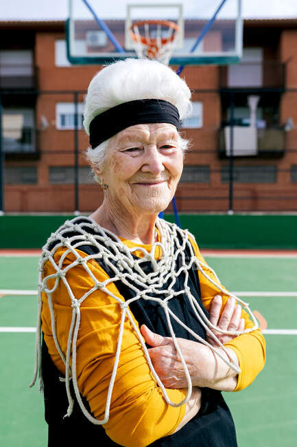 Sorrindo fêmea madura em activewear e rede branca olhando para a câmera enquanto em pé no chão de esportes com aro de basquete durante o treinamento — Fotografia de Stock
