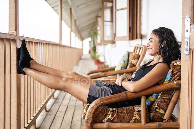 Vista lateral de la alegre joven turista étnica femenina en ropa casual sonriendo mientras se sienta en el sillón en la terraza de madera de la casa envejecida en el día soleado - foto de stock