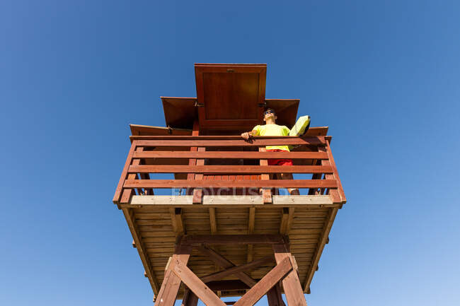 Angolo basso di coraggioso bagnino in occhiali da sole in piedi sulla torre di salvataggio in legno e monitoraggio della sicurezza sotto il cielo blu — Foto stock