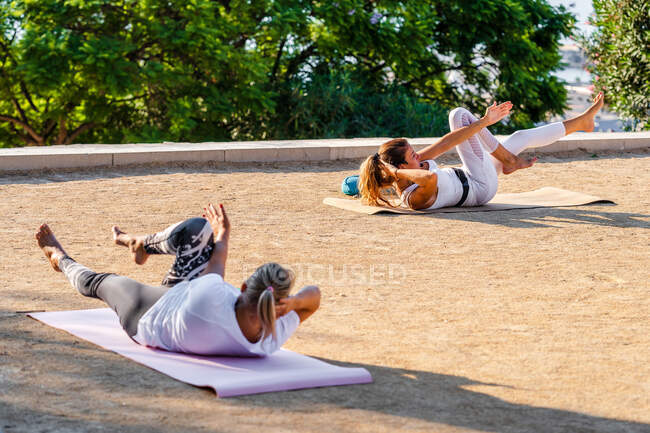 Спортивные дамы лежат на ковриках на террасе против деревьев и делают хрусты вместе при дневном свете — стоковое фото