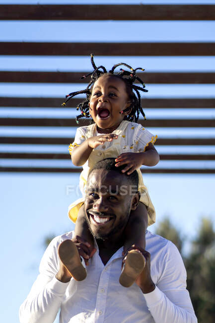 Счастливая афроамериканка с темными косичками, сидящая на плечах веселого отца и прыгающая, веселясь вместе на улице под солнцем — стоковое фото