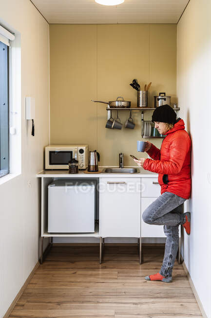 Вид сбоку на молодого путешественника в теплой куртке, стоящего на кухне, опираясь на стену, выпивая чашку горячего кофе и отправляя сообщения на мобильный телефон — стоковое фото