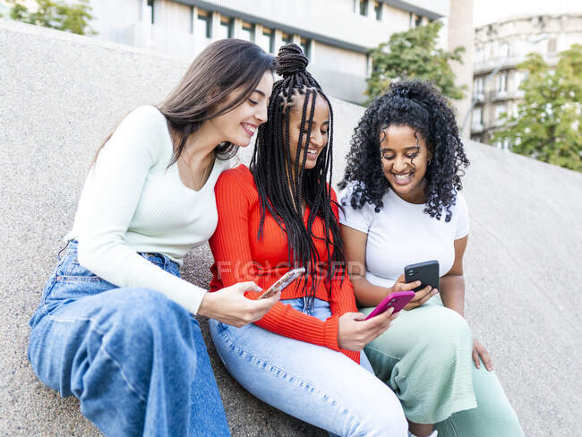 Cheerful multiracial girlfriends in trendy clothes laughing and showing video on smartphones on city street in daylight — Stock Photo