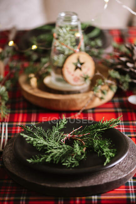 Cenário de mesa de Natal com coroa de flores e ornamentos decorativos de madeira e toalha de mesa quadriculada vermelha com luzes amarelas no fundo — Fotografia de Stock
