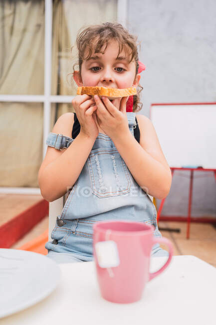 Carino ragazza in denim generale guardando verso il basso mentre si mangia pane fresco con marmellata dolce nella stanza luce a casa — Foto stock