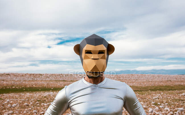Homme en masque de singe et tenue en latex argenté debout sur un champ pierreux et regardant la caméra contre un ciel bleu nuageux — Photo de stock