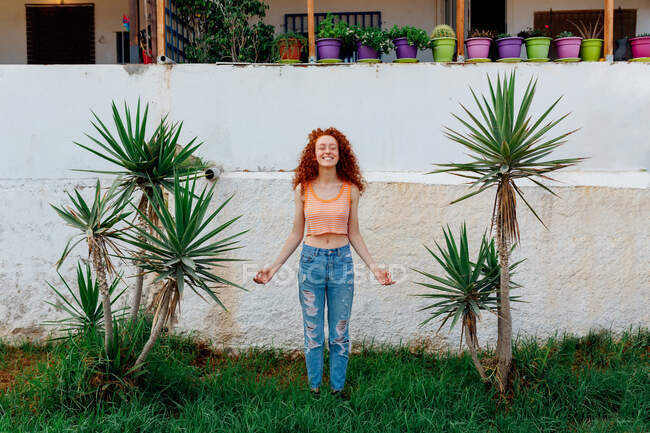 Full body of cheerful female with long curly hair making zen gesture while meditating between exotic palms — Stock Photo