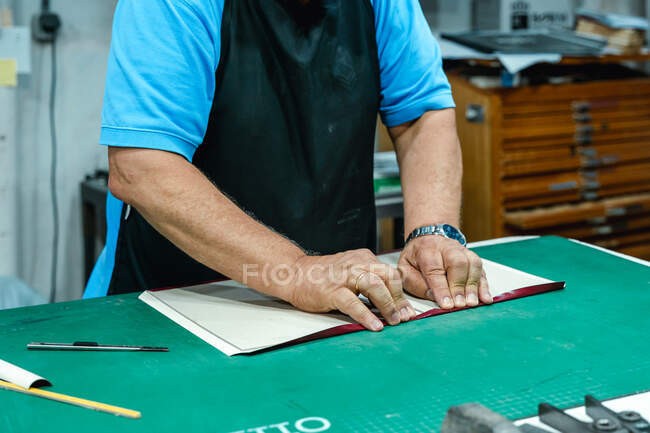 Crop unrecognizable male artisan in casual clothes and apron folding cover of album while standing at workbench in traditional printing studio — Stock Photo