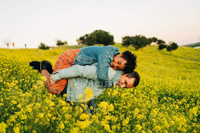 Jovem romântico sorrindo e dando passeio de piggyback para a alegre namorada afro-americana no exuberante prado amarelo florescendo no campo — Fotografia de Stock