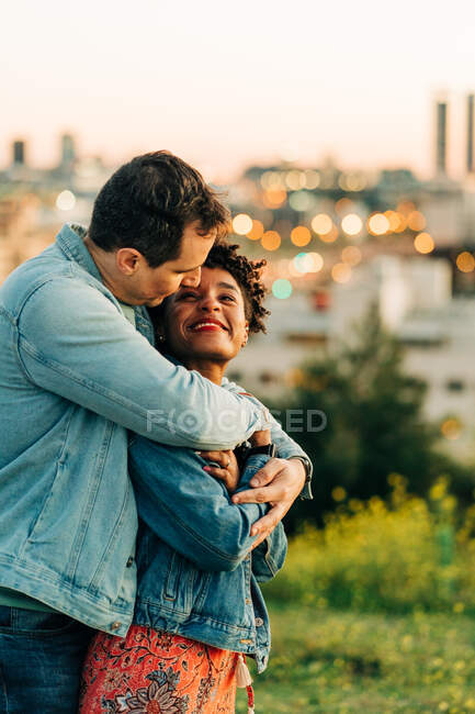 Romántica pareja diversa abrazándose y mirándose mientras están de pie en el césped contra el paisaje urbano con edificios en un fondo borroso - foto de stock