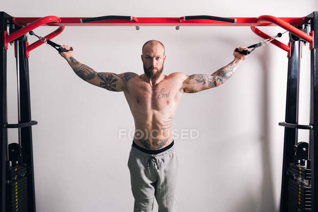 Muskelkräftiger, hemdsloser Bodybuilder, der im Fitnessstudio steht und Übungen auf einem Seil-Crossover-Gerät macht und in die Kamera schaut — Stockfoto