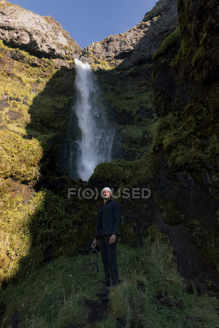 Low angle of attentive young Hispanic male traveler in warm clothes and hat looking away while standing near picturesque waterfall flowing through rocky ravine with photo camera in hands — Stock Photo