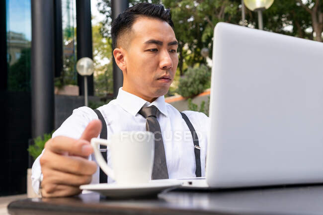 Жалкий молодой азиатский предприниматель с чашкой горячего напитка и нетбуком, смотрящий на экран в городском кафетерии при дневном свете — стоковое фото