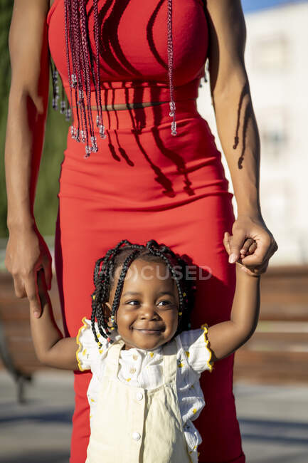 Feliz niña afroamericana en vestido ligero cogida de la mano de una madre irreconocible mientras camina por la calle en un día soleado - foto de stock