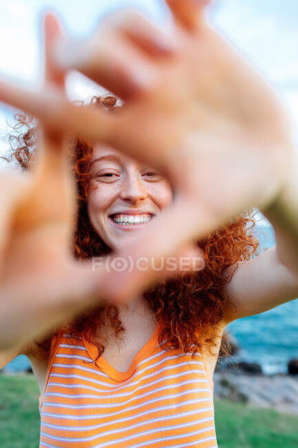 Mulher positiva com longos cabelos encaracolados de gengibre moldando triângulo na praia com pedras e olhando para a câmera — Fotografia de Stock