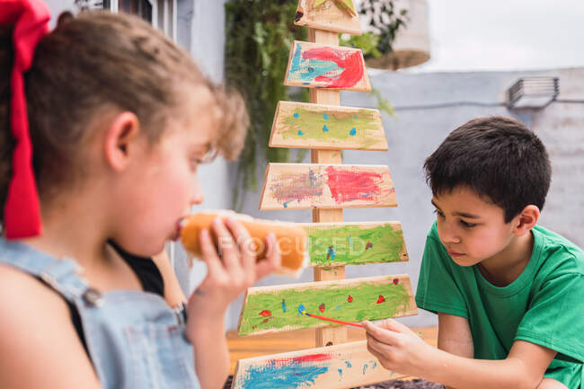 Fille manger hot dog près de garçon peinture arbre de Noël décoratif avec pinceau dans la pièce lumineuse pendant la préparation des vacances à la maison — Photo de stock