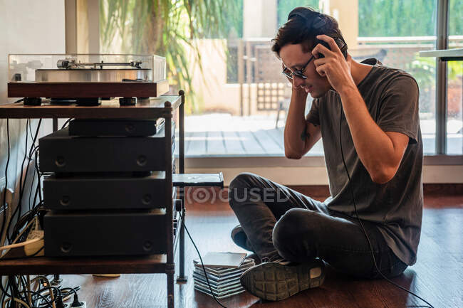Vista laterale dell'uomo in cuffia che ascolta musica mentre è seduto sul pavimento vicino al lettore a casa — Foto stock