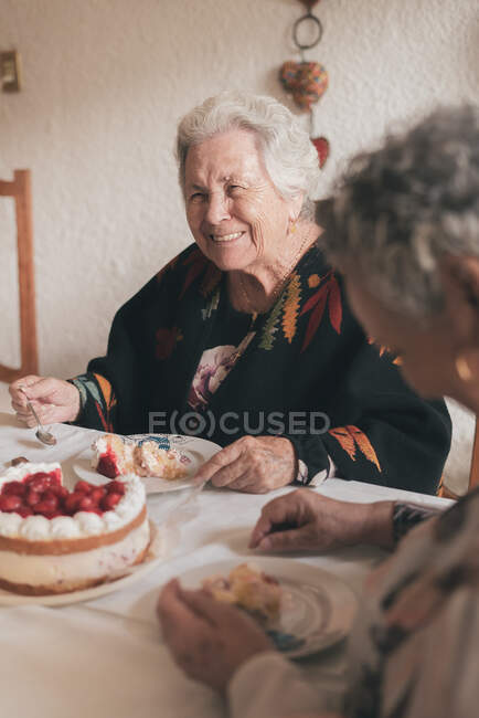 Mulher idosa com cabelos grisalhos e fêmea sênior sentada à mesa de jantar e comemorando 90 anos com delicioso bolo com velas — Fotografia de Stock
