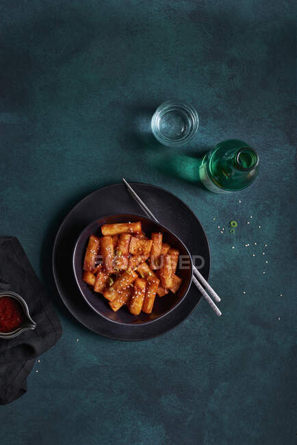 Pile of stir fried spicy rice cakes tteokbokki topped with sesame seeds served in black bowl on table with alcohol drink — Stock Photo